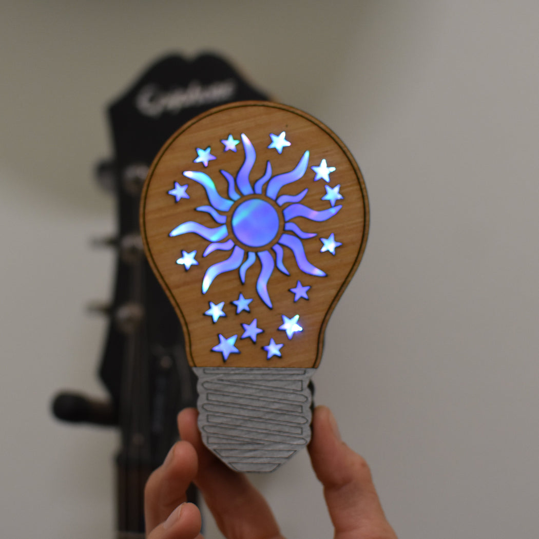 Hand holding a wooden light bulb-shaped object with sun and star design against a blurred background